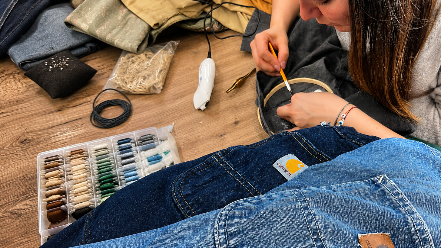 A person sits on a wooden floor surrounded by sewing and crafting supplies, working on a circular embroidery or knitting project. In the foreground, a pair of blue denim jeans fills much of the frame, with a small yellow logo tab visible on the pocket.